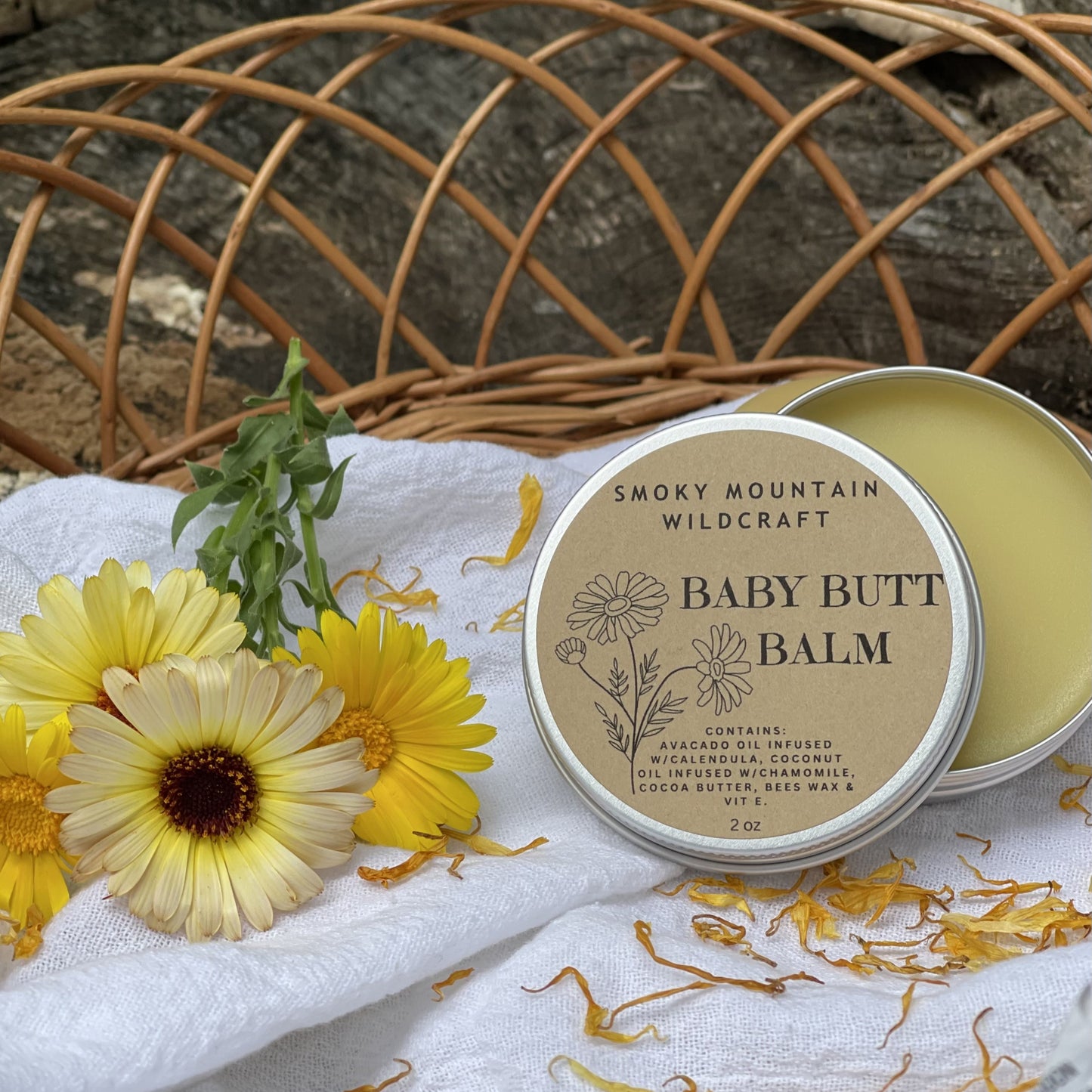 Silver tin of a product "Baby Butt Balm" made by smoky mountain wildcraft. Sitting slightly open in a woven basket with calendula flowers around it.