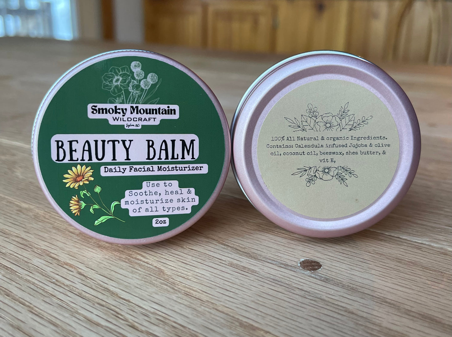 front and back shot of a pink 2 oz circle tin of a product called beauty balm a all natural facial lotion made by Smoky Mountain WIldcraft. The tin has a green label against a wooden background.