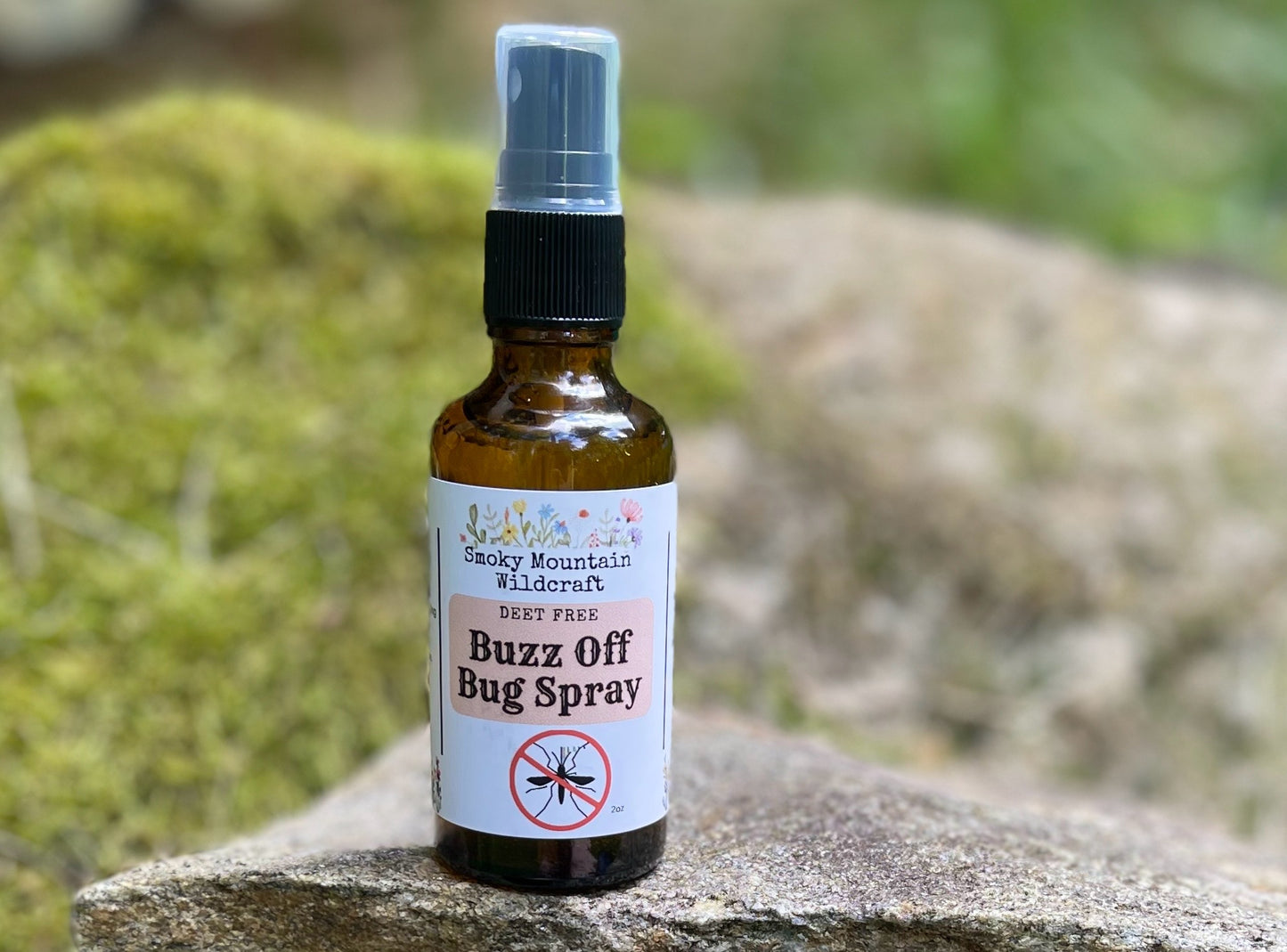 An amber spray bottle of 'Buzz Off Bug Spray' by Smoky Mountain Wildcraft, featuring an all-natural formula of essential oils positioned on a rock with a green, natural background.