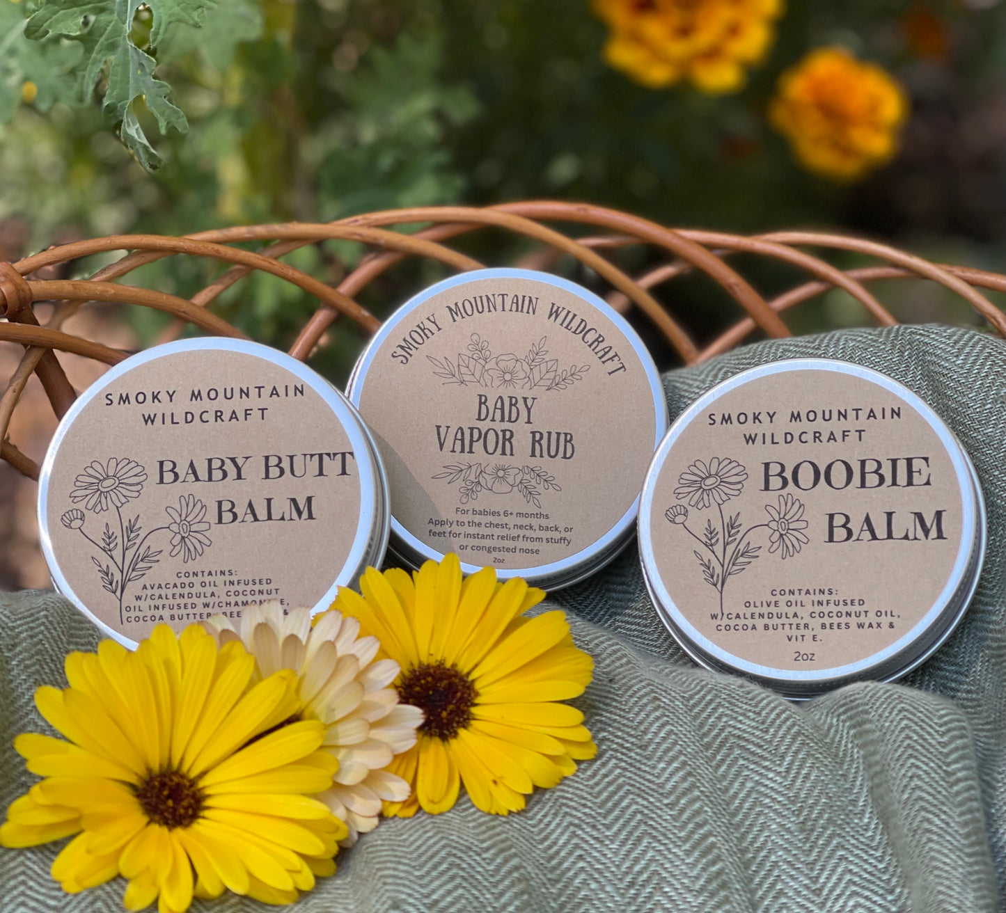 3 silver tins of an all natural, organic salve for babies. Made by Smoky Mountain WIldcraft. The tins are labeled "baby butt balm", "Baby vapor rub" and "Boobie balm" Sitting in a woven basket with yellow calendula flowers sitting around the tins.