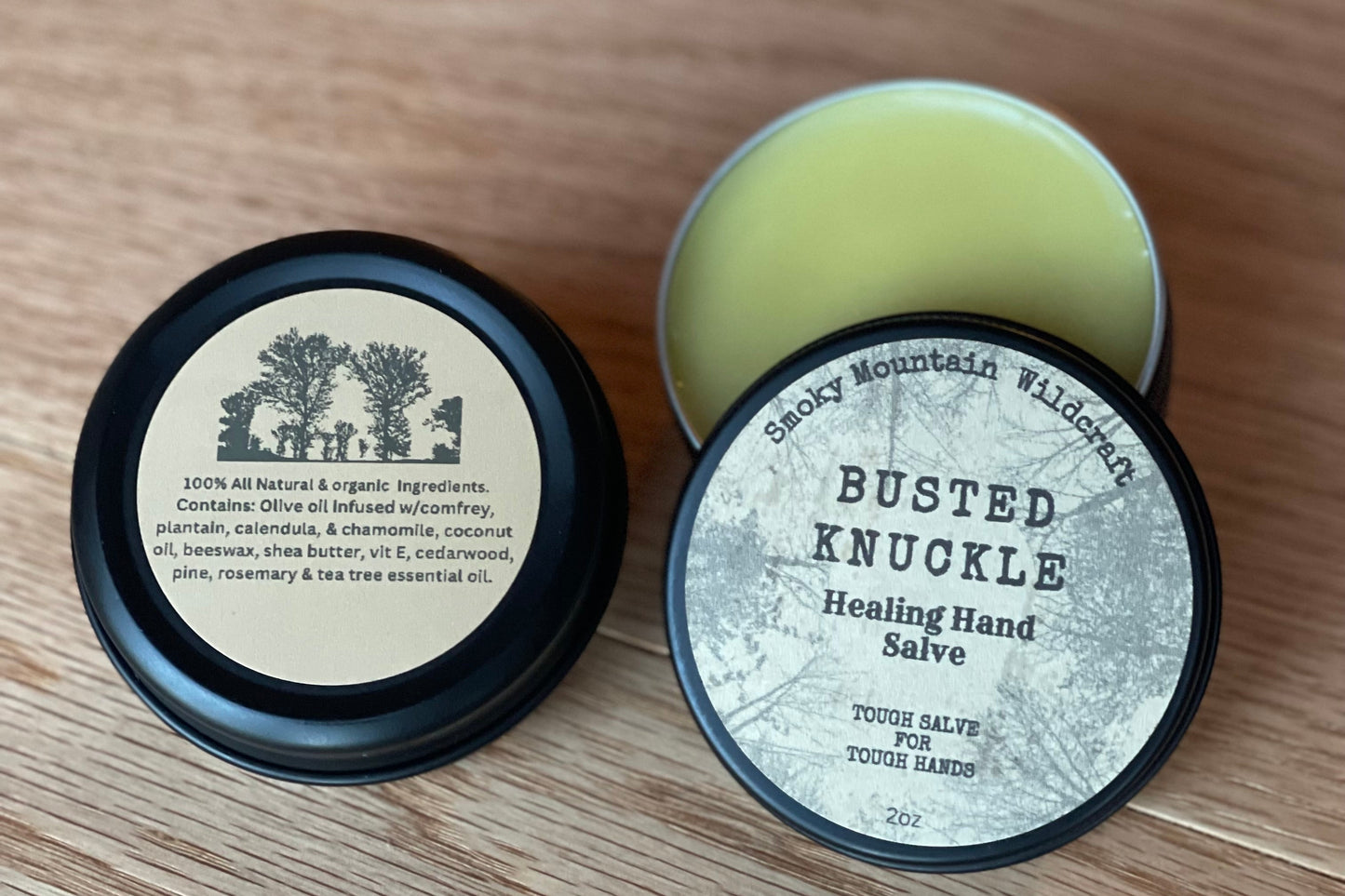 two circle black tins of a product called "busted knuckle healing hand salve" One tin is open to show the inside yellow salve and the other tin is a view of the back of the tin with all of the organic ingredients.