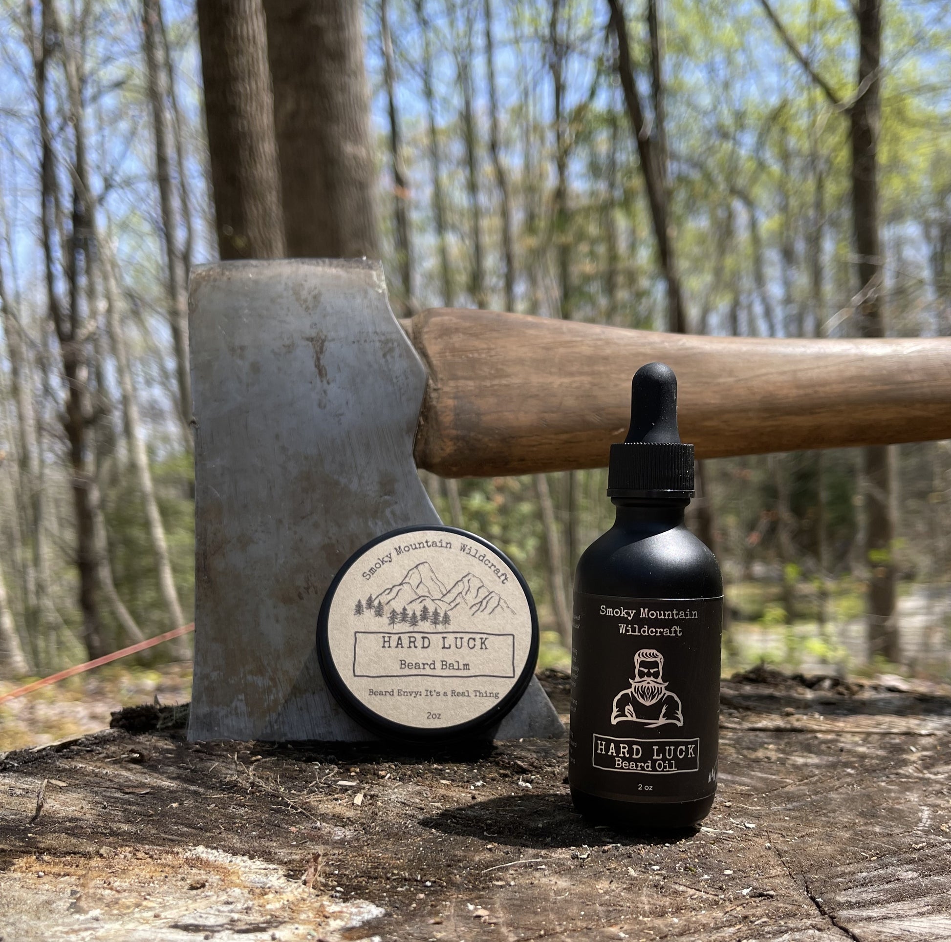 A bottle of 'Hard Luck' beard oil and a tin of a beard balm. products on a wooden surface with a axe in the background.