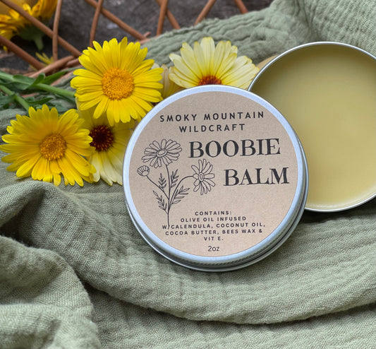 Silver circle tin open to show yellow salve. Sitting in a woven basket on a green blanket with calendula flower around it. 
