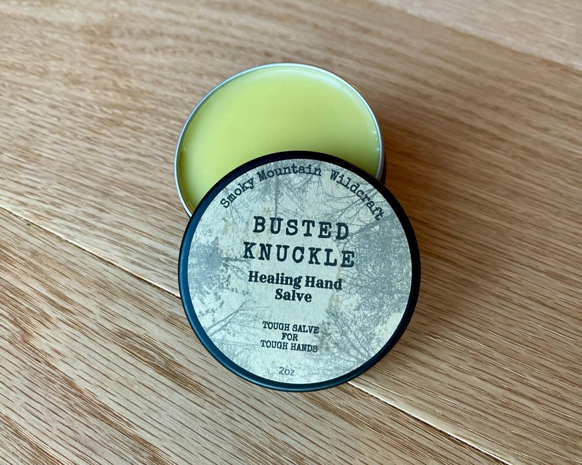 A container of Busted Knuckle Healing Hand Salve with a vintage-style label, placed on a wooden surface. In a black circle tin that is partially open so that you can see the golden hand salve.

Details