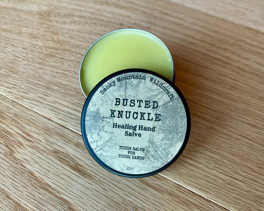 A container of Busted Knuckle Healing Hand Salve with a vintage-style label, placed on a wooden surface. In a black circle tin that is partially open so that you can see the golden hand salve.

Details