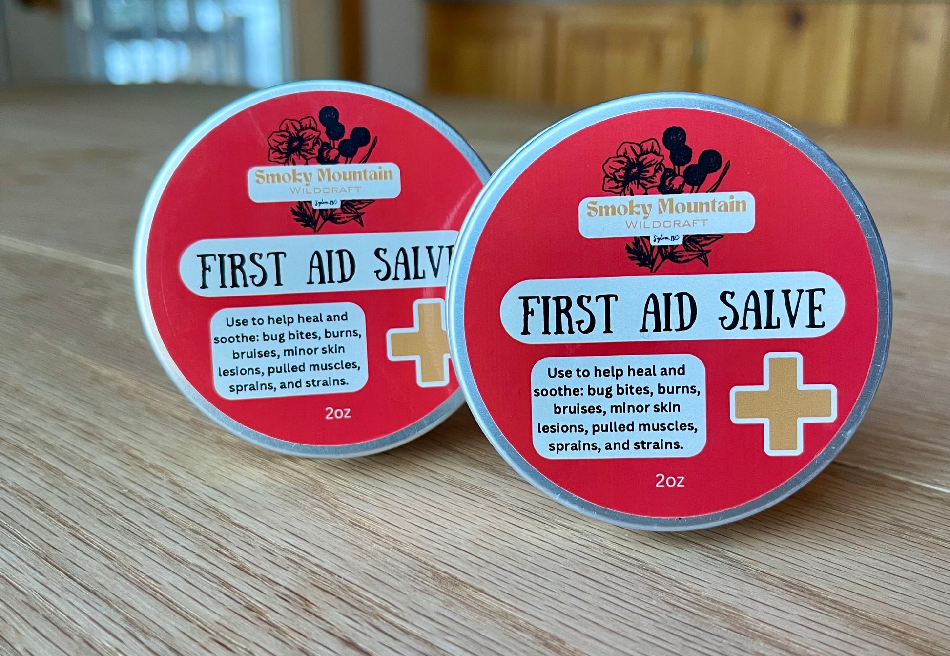 Two tins of First Aid Salve with red labels, one behind the other, on a wooden surface. The front of the tins display the product name and list some of its ingredients.