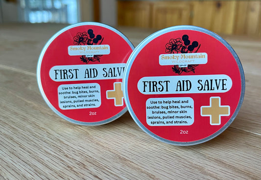 Two tins of First Aid Salve with red labels, one behind the other, on a wooden surface. The front of the tins display the product name and list some of its ingredients.