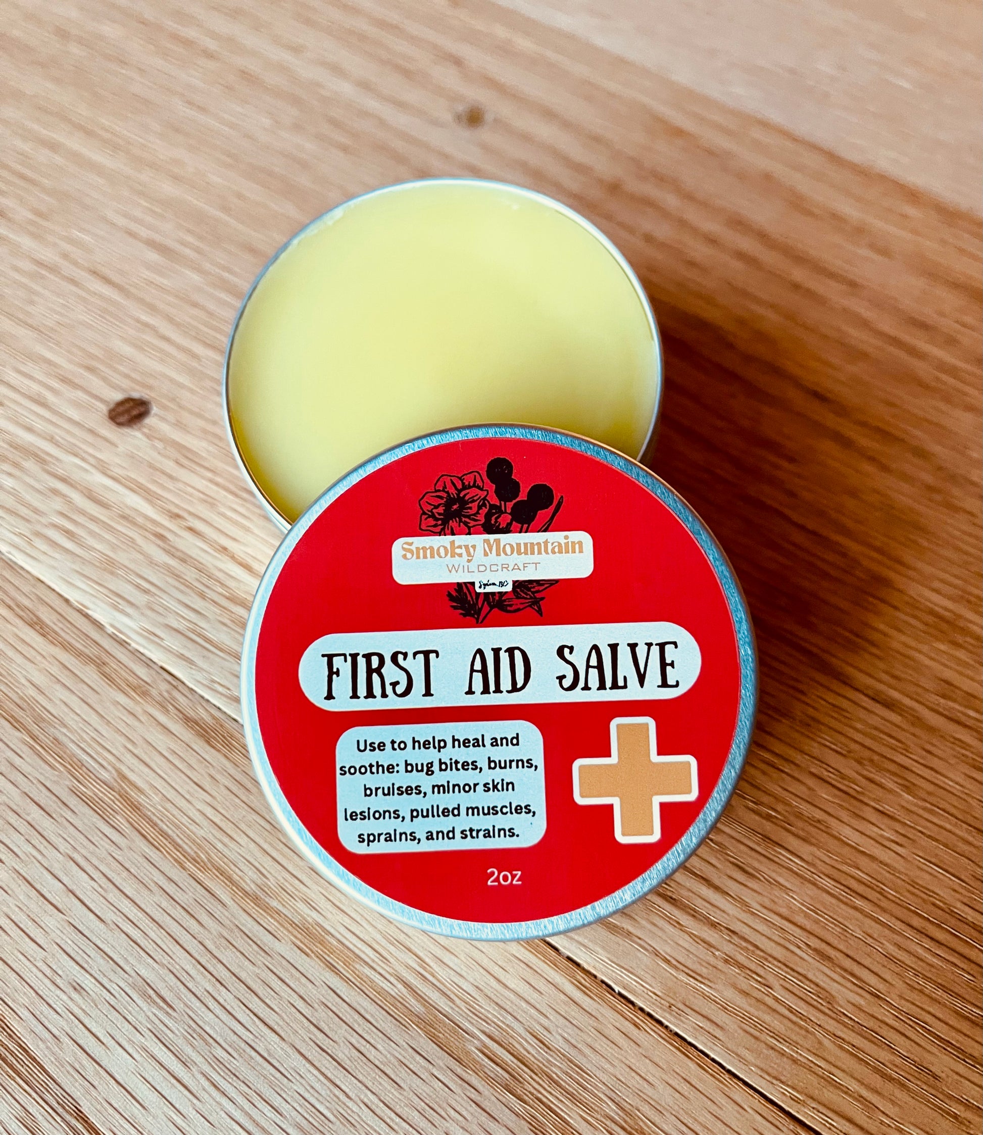 Open tins of First Aid Salve with a red label. Tine is open showing the golden yellow salve and it is sitting on a wooden surface. The front of the tins display the product name and use.