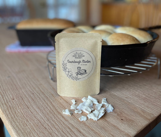 A packet of Sourdough Starter next to a loaf of bread in a pan, with more bread in the background, all on a wooden table.

