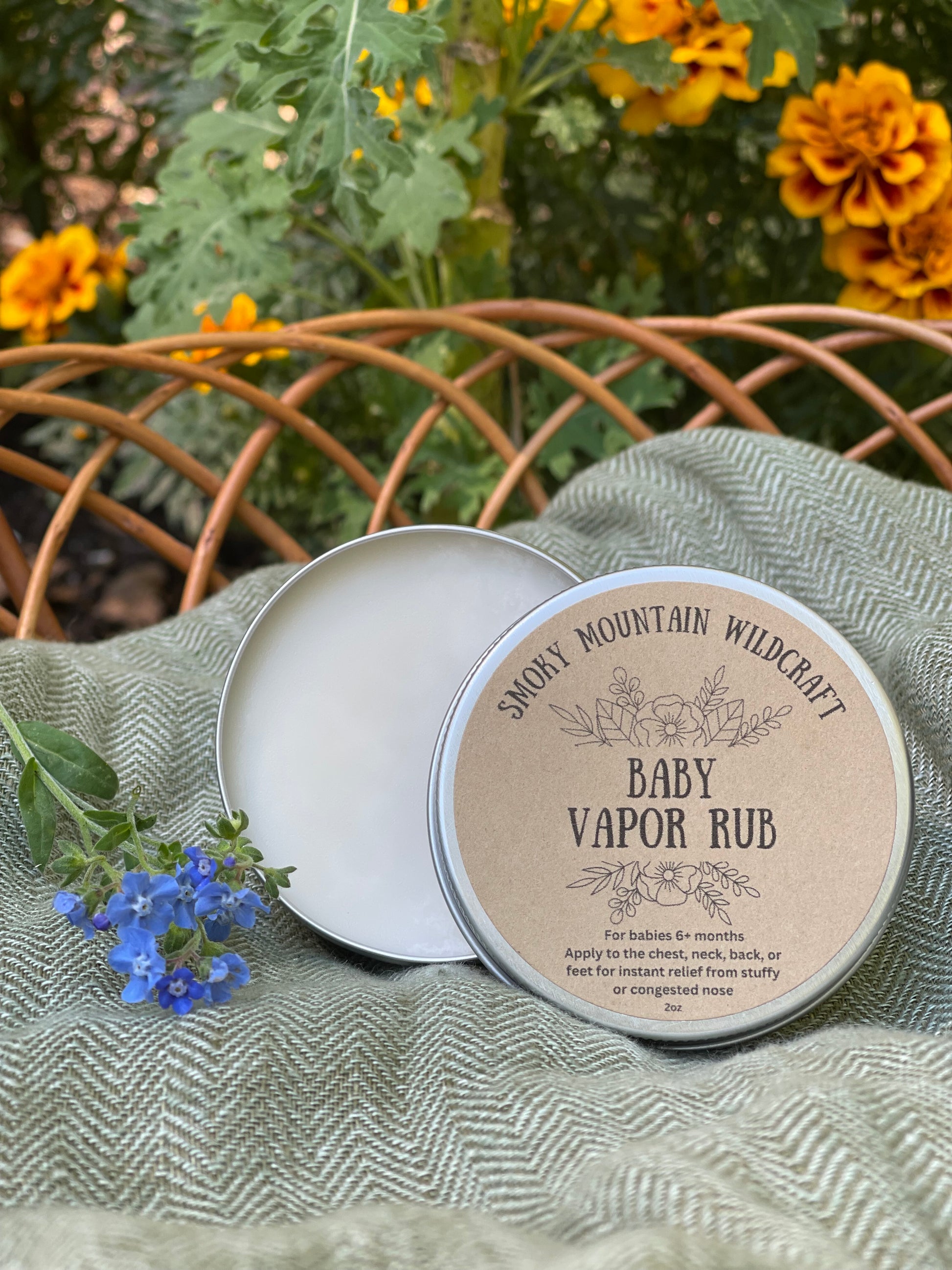 Silver 2 oz can of 'Baby Vapor Rub' by Smoky Mountain Wildcraft, placed on a fabric with flowers, indicating herbal nature of the product