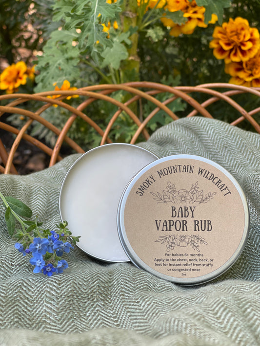 Silver 2 oz can of 'Baby Vapor Rub' by Smoky Mountain Wildcraft, placed on a fabric with flowers, indicating herbal nature of the product