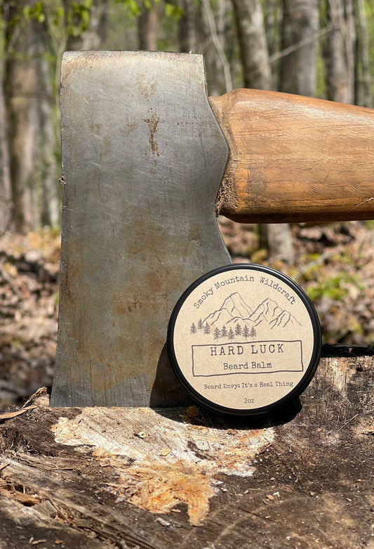 Black circle tin of 'Hard Luck Beard Balm ' product on a log in a forest setting with a axe in the log in the background.
