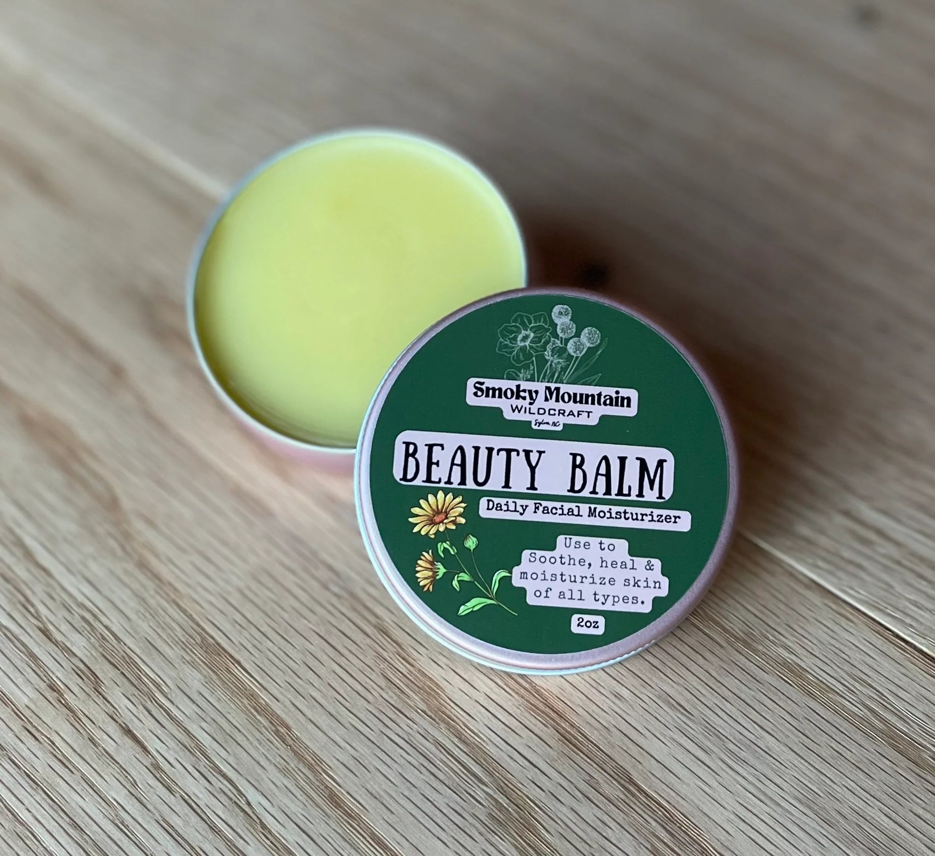 pink 2 oz circle tin of a product called beauty balm  a all natural facial lotion made by Smoky Mountain WIldcraft. The tin has a green label against a wooden background.