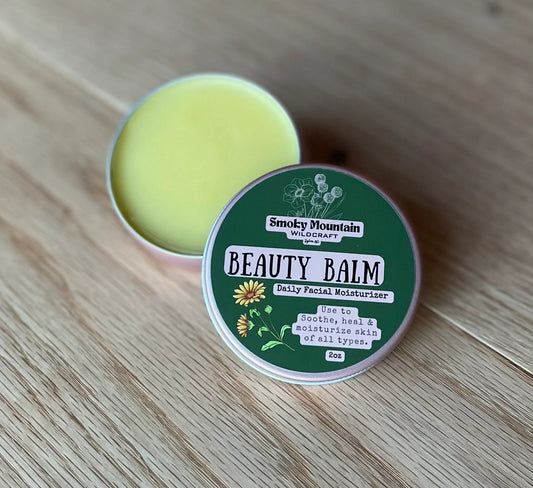 pink 2 oz circle tin of a product called beauty balm  a all natural facial lotion made by Smoky Mountain WIldcraft. The tin has a green label against a wooden background.