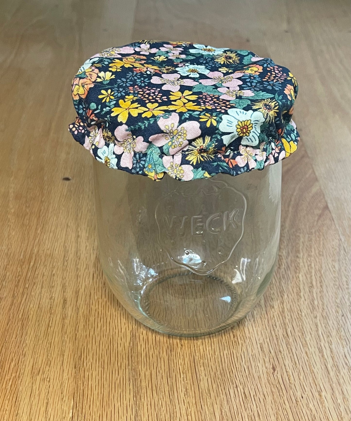Clear glass jar with a floral-patterned jar cover on a wooden surface. Jar cover is meant for sourdough or sourdough starter.

