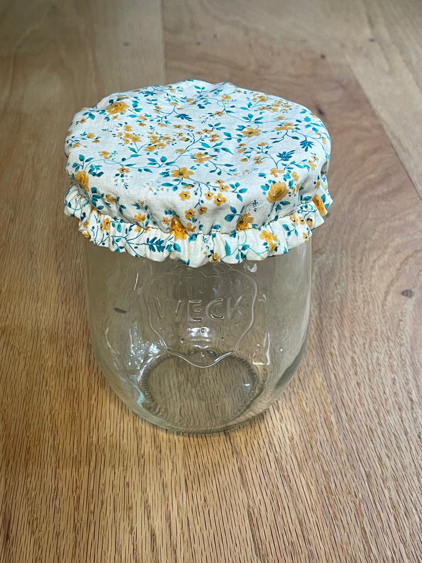 Floral-patterned fabric on a clear plastic container on a wooden surface. Jar cover is meant for sourdough or sourdough starter.